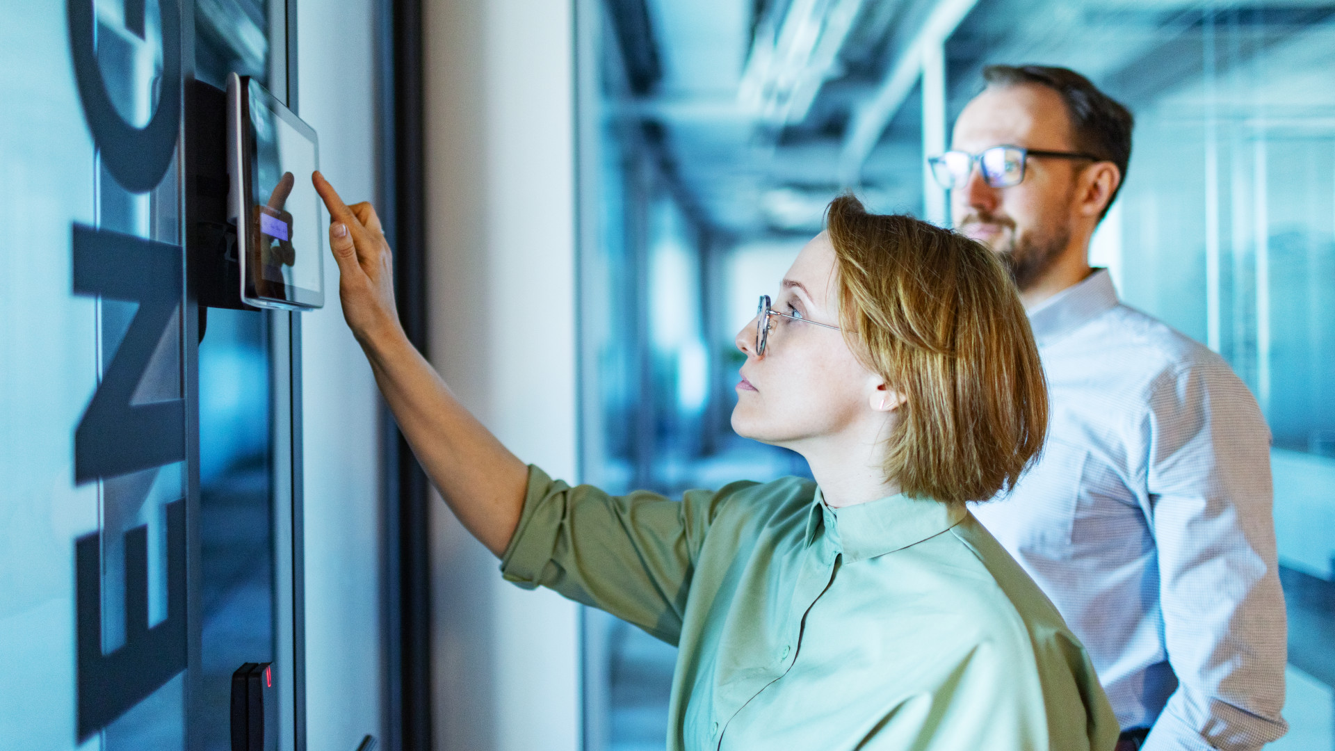 Eine Frau bedient ein Touchpad eines Smart Offices und im Hintergrund steht ein Kollege